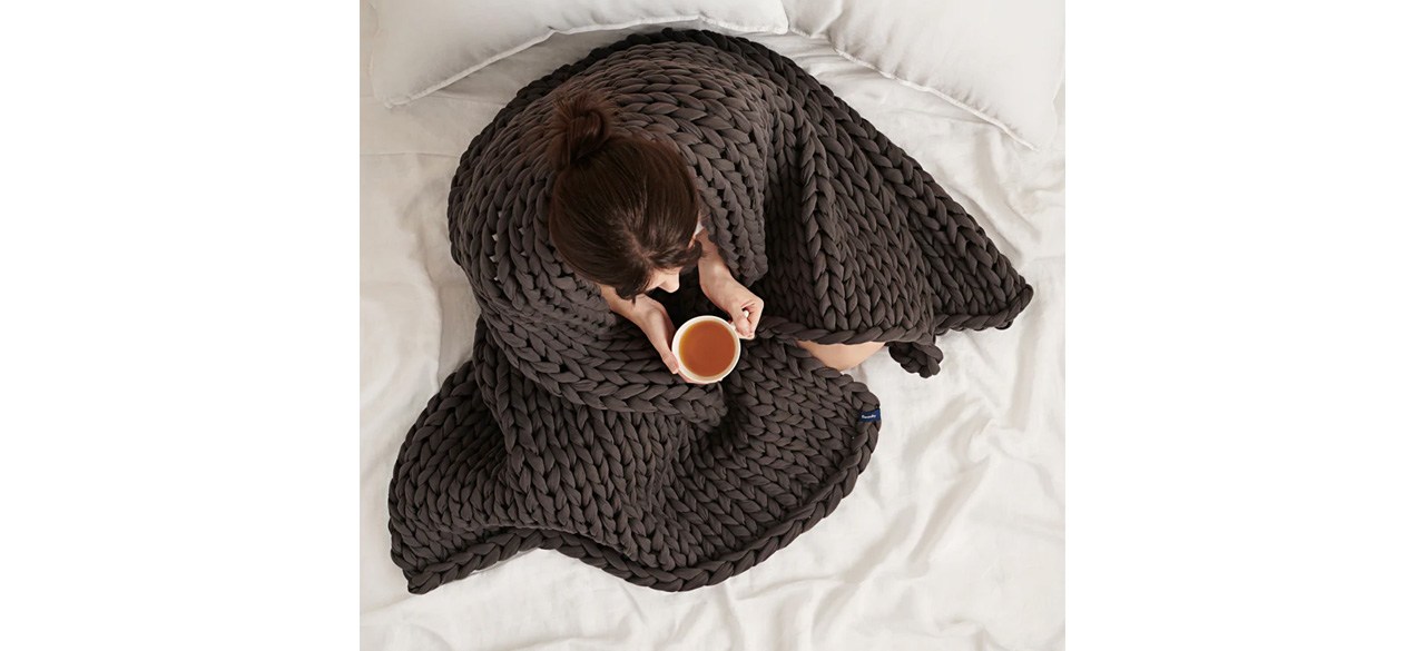 A person wearing the Bearaby Cotton Napper Weighted Blanket and holding a warm beverage