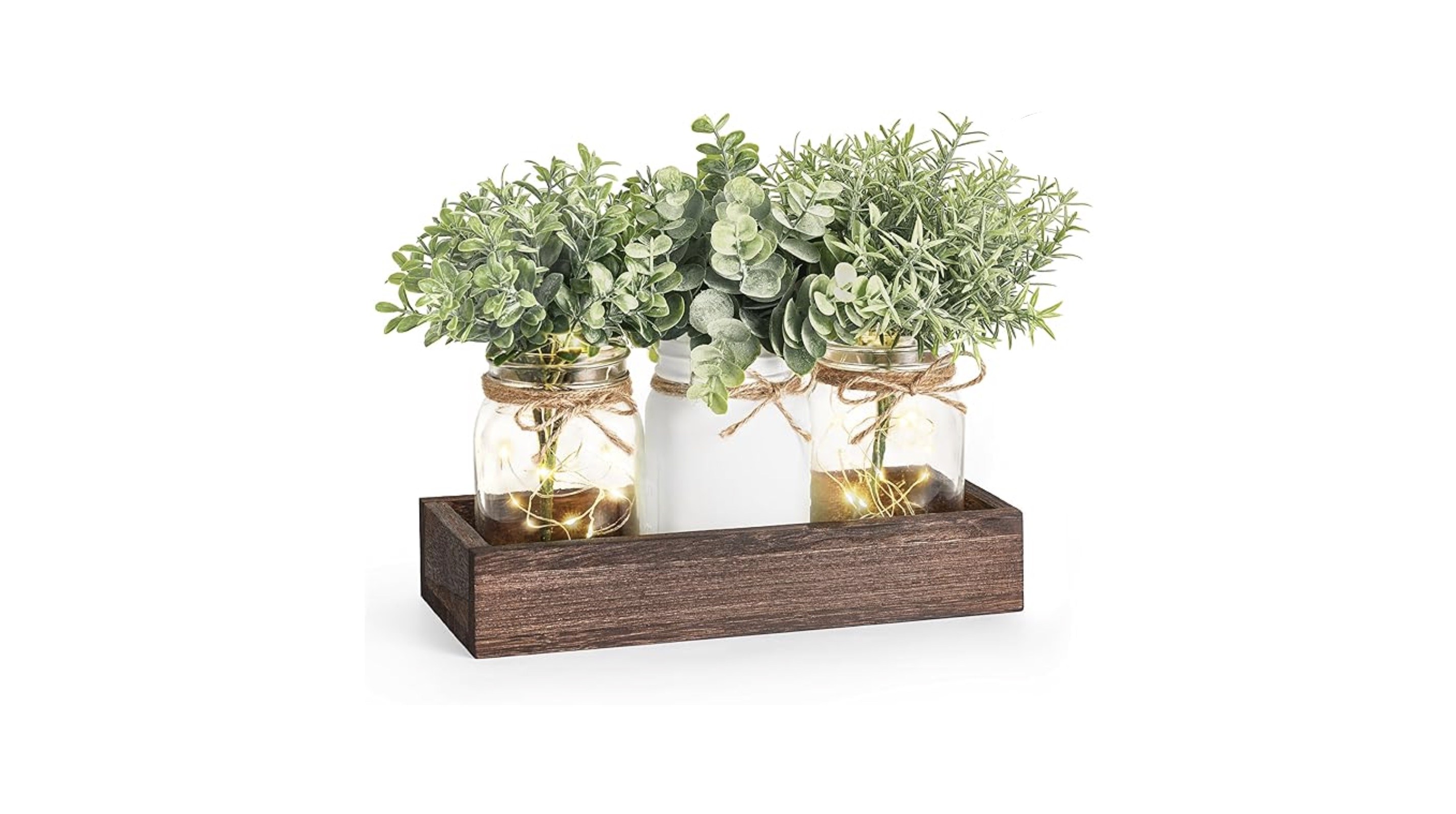 A wooden centerpiece with 3 jars on it: one white in the middle and 2 clear ones with fairy lights in them on the sides. All are holding plants.